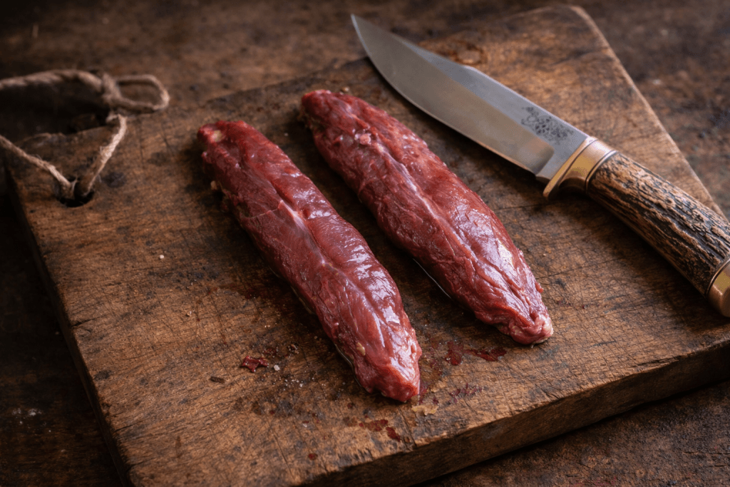 Mușchii de căprior (mușchii popii) proaspăt extrași, așezați pe un tocător rustic de lemn lângă un cuțit de vânătoare.
